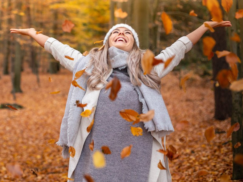 Life Extension, happy and vital woman in the woods with arms stretched out surrounded with orange fall leaves around her.